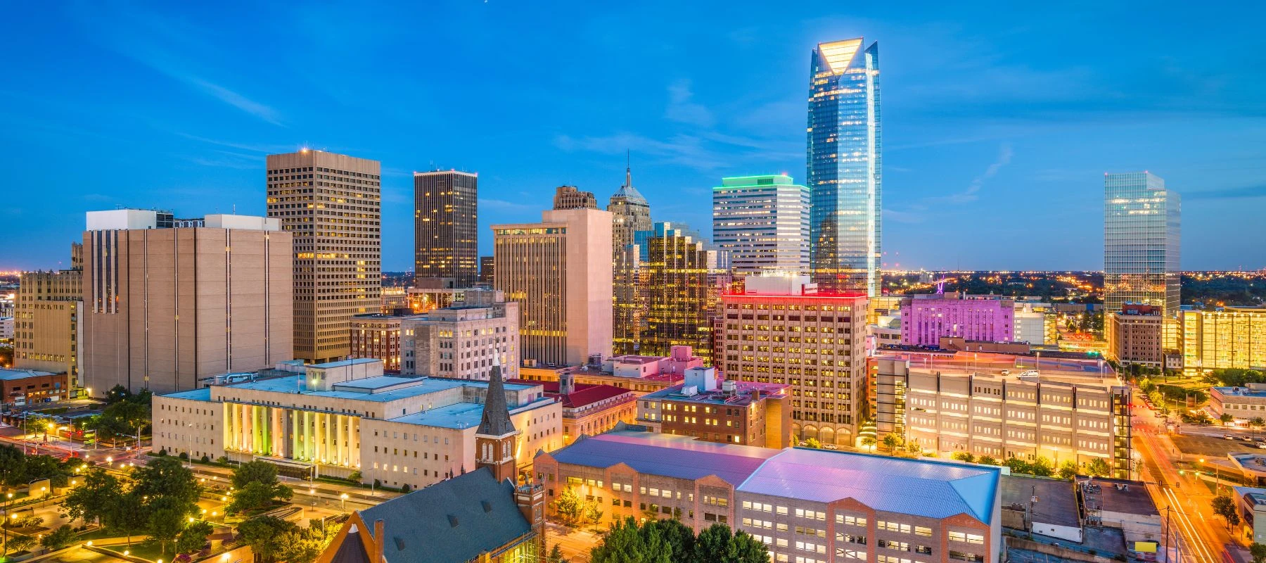 Downtown city skyline at dusk with modern high-rise buildings, colorful lights illuminating the structures, and a clear blue sky in the background. Streets and trees are visible in the foreground.