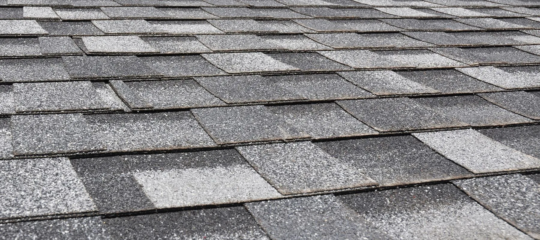 Close-up view of an asphalt shingle roof with overlapping gray and white rectangular shingles in a repeating pattern. The texture shows granules on the surface of each shingle.