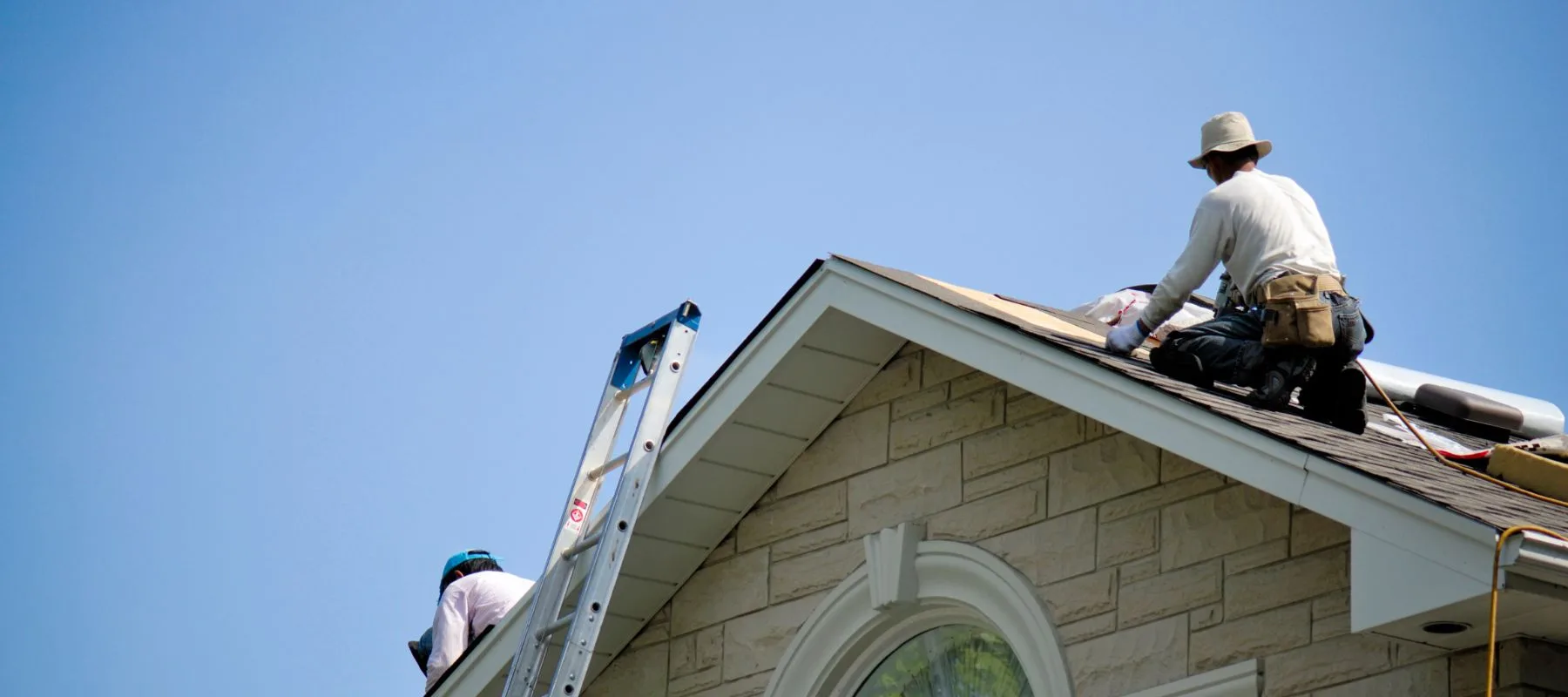 Two workers on the roof of a house, one near a ladder and the other working with roofing materials, both wearing hats and safety gear under a clear blue sky.