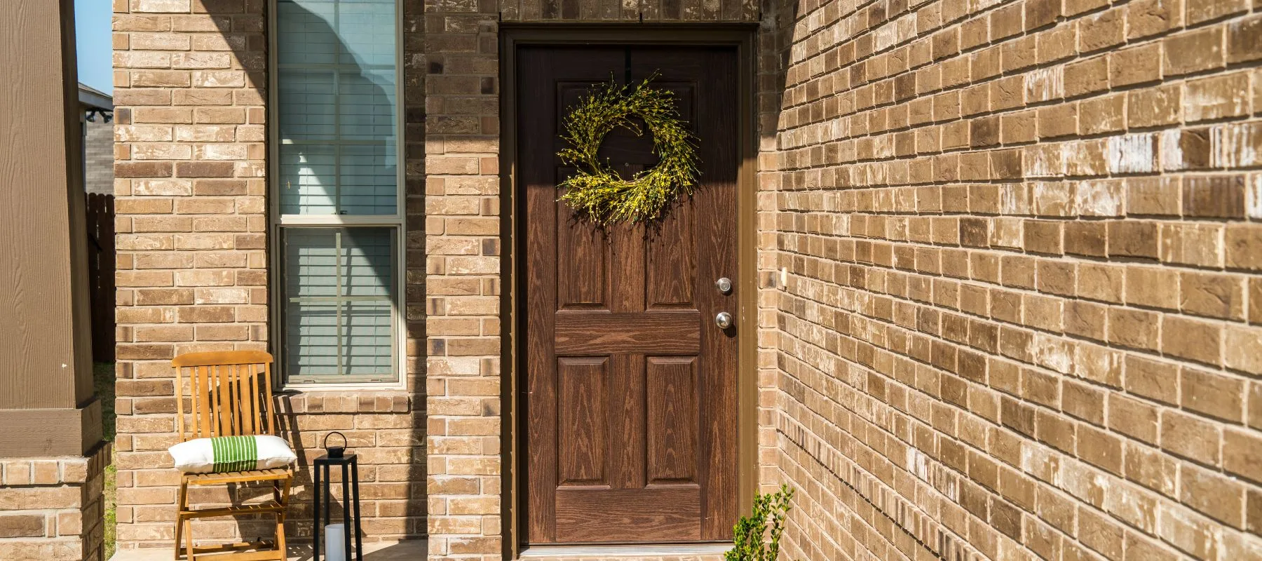 A brown wooden front door with a wreath, set in a brick house. A wooden chair with a cushion and a black lantern sit to the left of the door near a window. Bright sunlight casts shadows on the scene.