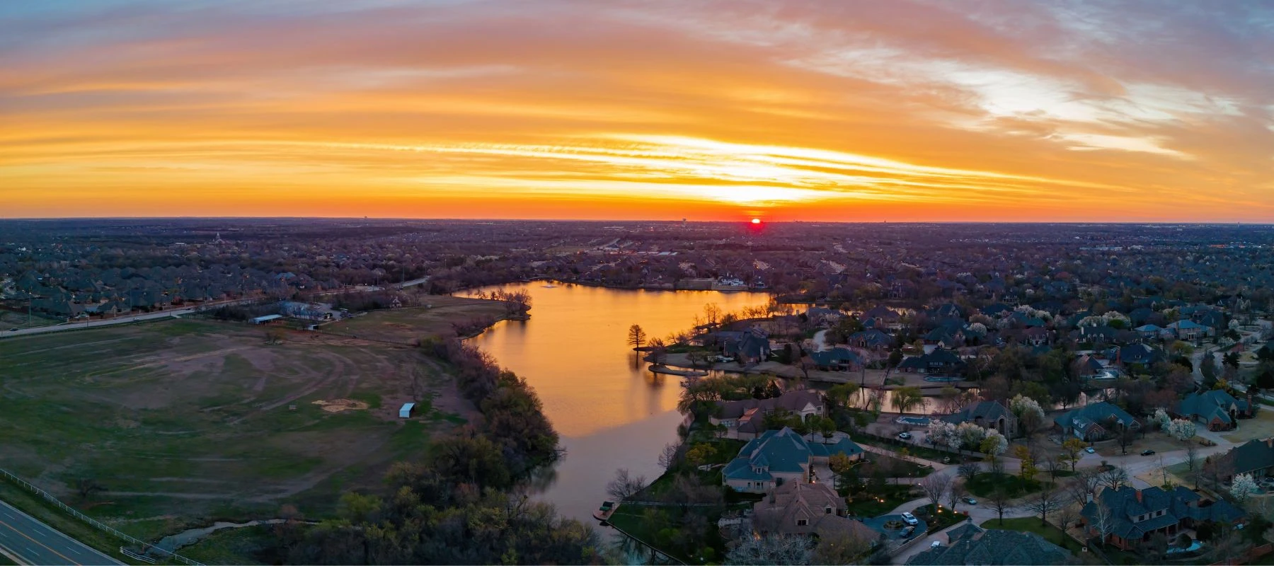 Aerial view of a suburban neighborhood with houses surrounding a reflective lake at sunset, vibrant orange and yellow hues fill the sky, casting a warm glow over the landscape.