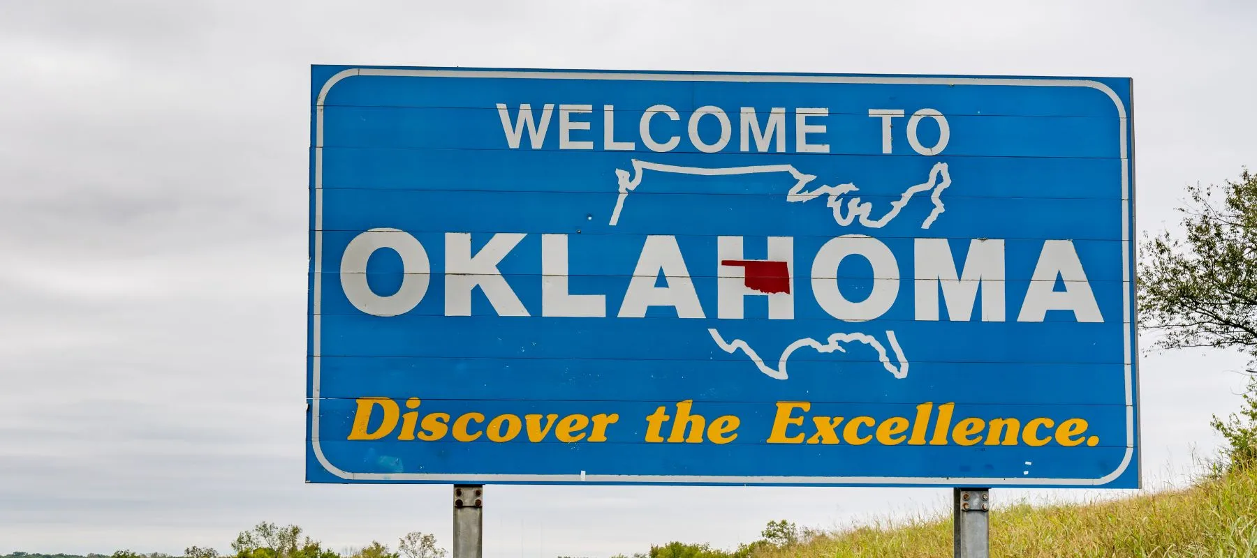 Blue roadside sign reads Welcome to Oklahoma with an outline of the U.S. and a red box marking Oklahoma, and the slogan Discover the Excellence. Some grass and trees are visible nearby.