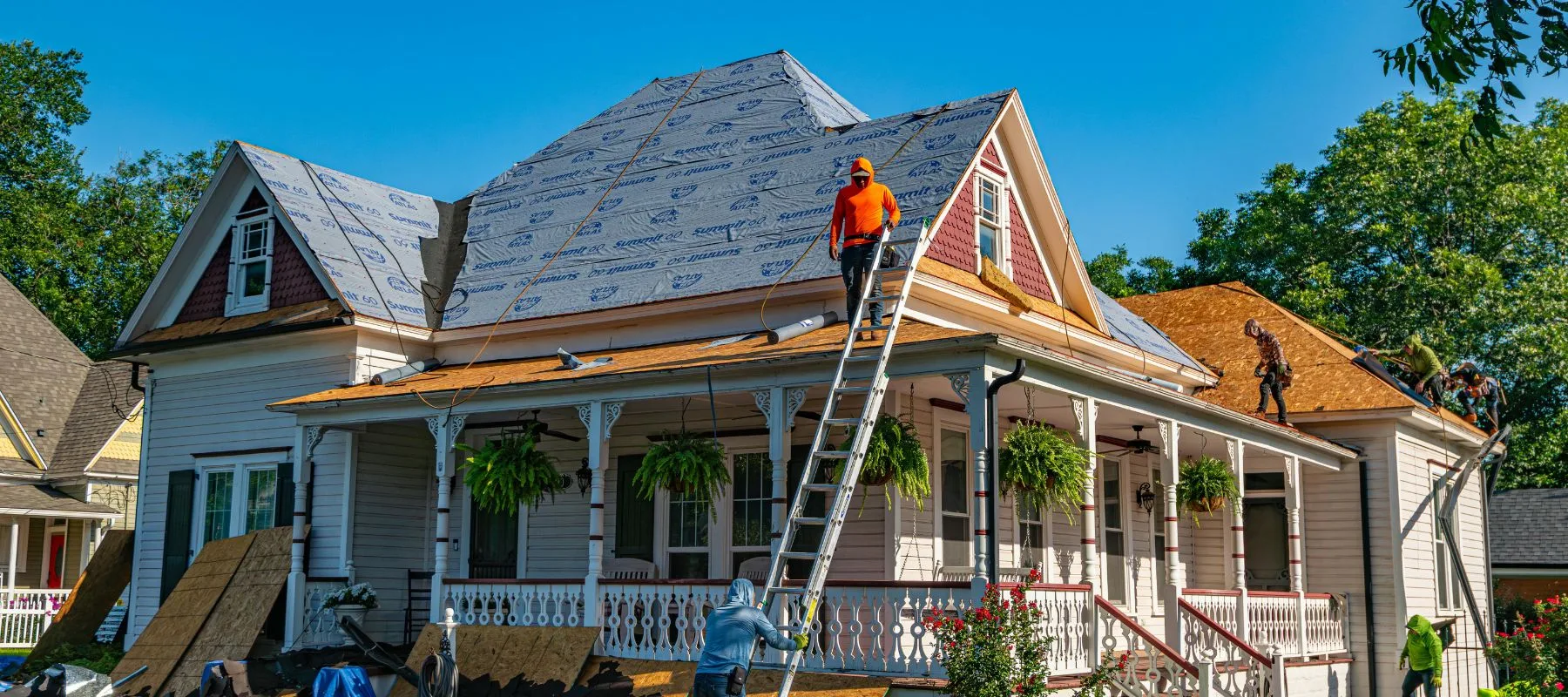 Workers repair the roof of a large white house, with one person on a ladder and others on the roof. The roof is partially covered with underlayment. Tools and building materials are visible around the house.
