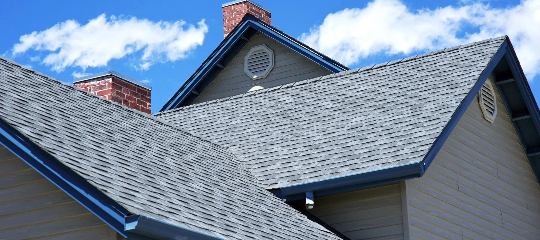 Close-up view of a house roof with gray asphalt shingles, blue trim, two brick chimneys, and a triangular attic vent, set against a bright blue sky with scattered white clouds.