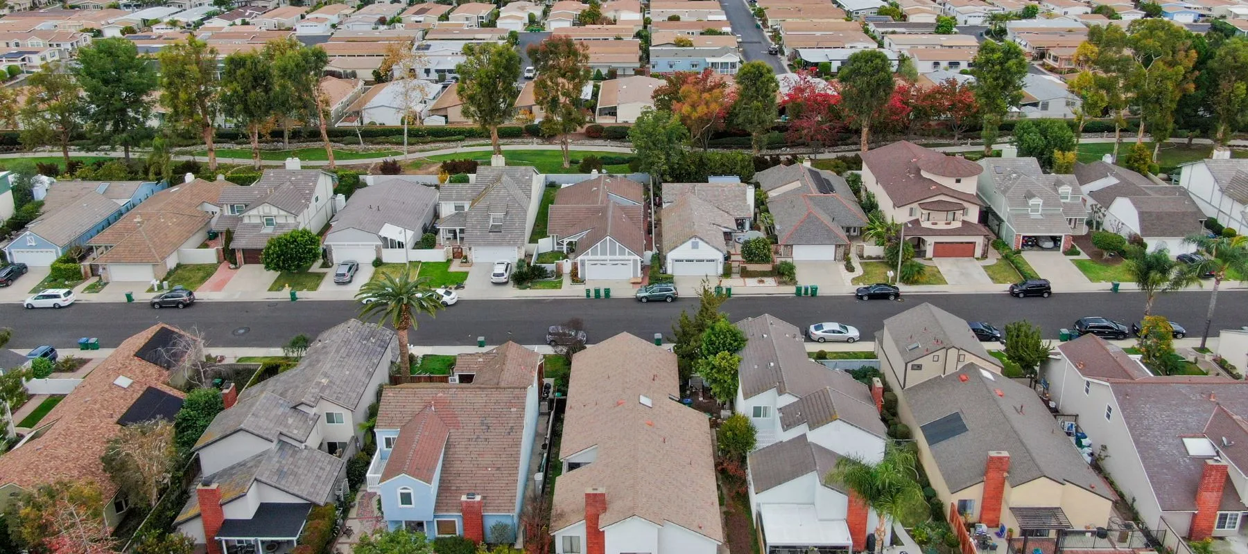 Aerial view of a suburban neighborhood with rows of houses, tree-lined streets, parked cars, green lawns, and a grid-like street layout.