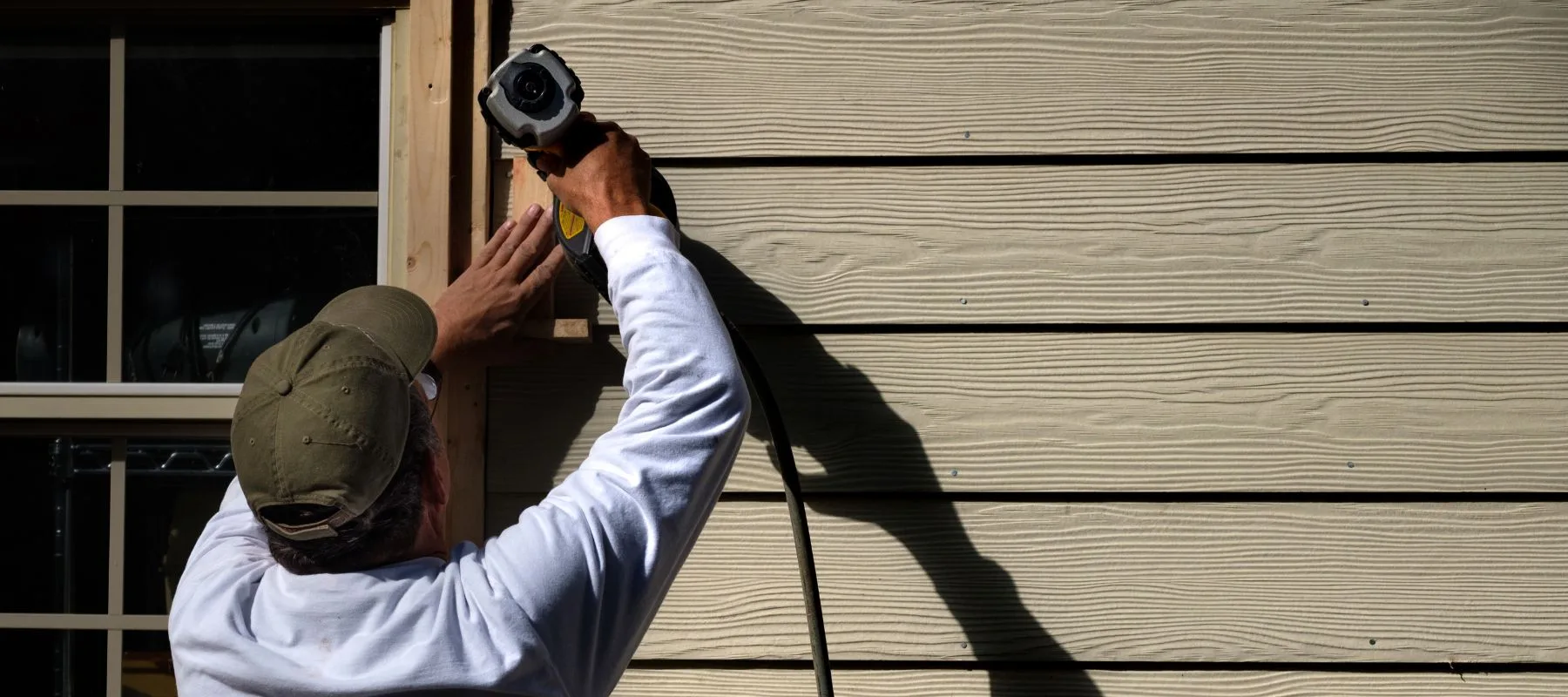 A person wearing a cap and white shirt uses a nail gun to install siding on a house, casting a shadow on the wall in bright sunlight.