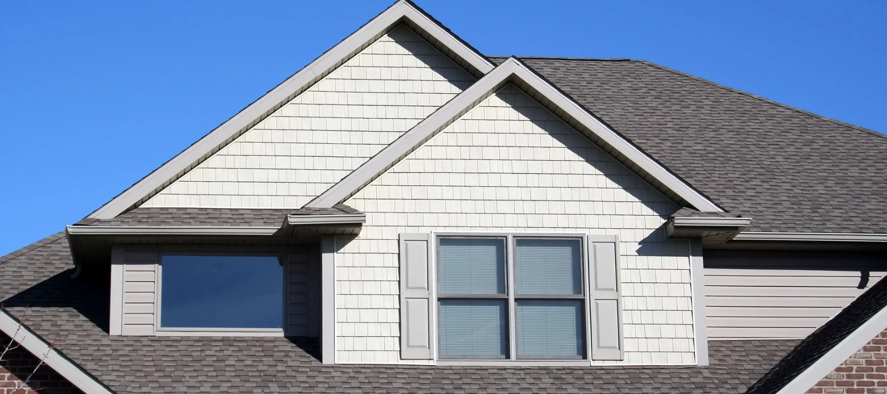 The upper portion of a house with light-colored siding, two gables, gray asphalt shingles, a set of double windows with shutters, and a single window, all under a clear blue sky.