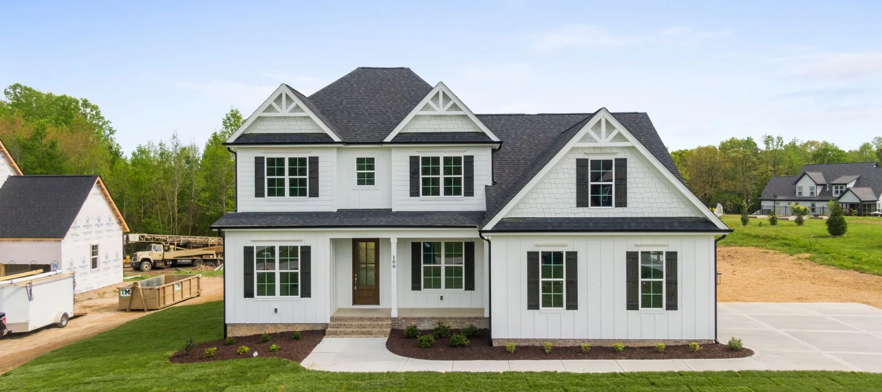 A modern two-story white house with dark roof shingles, multiple gables, large windows, a covered front porch, and a concrete driveway, surrounded by green grass and trees under a clear sky.